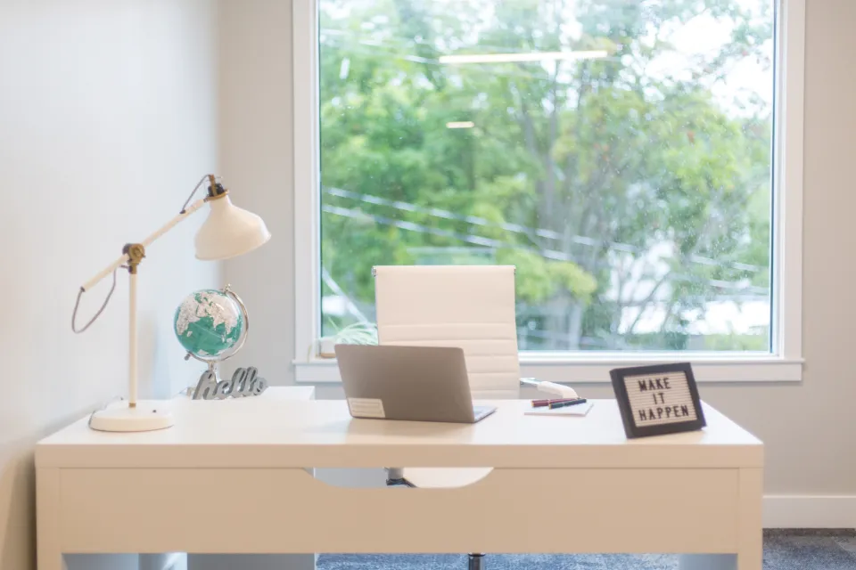 A tidy white desk by a bright window, with a laptop, a desk lamp and a small globe