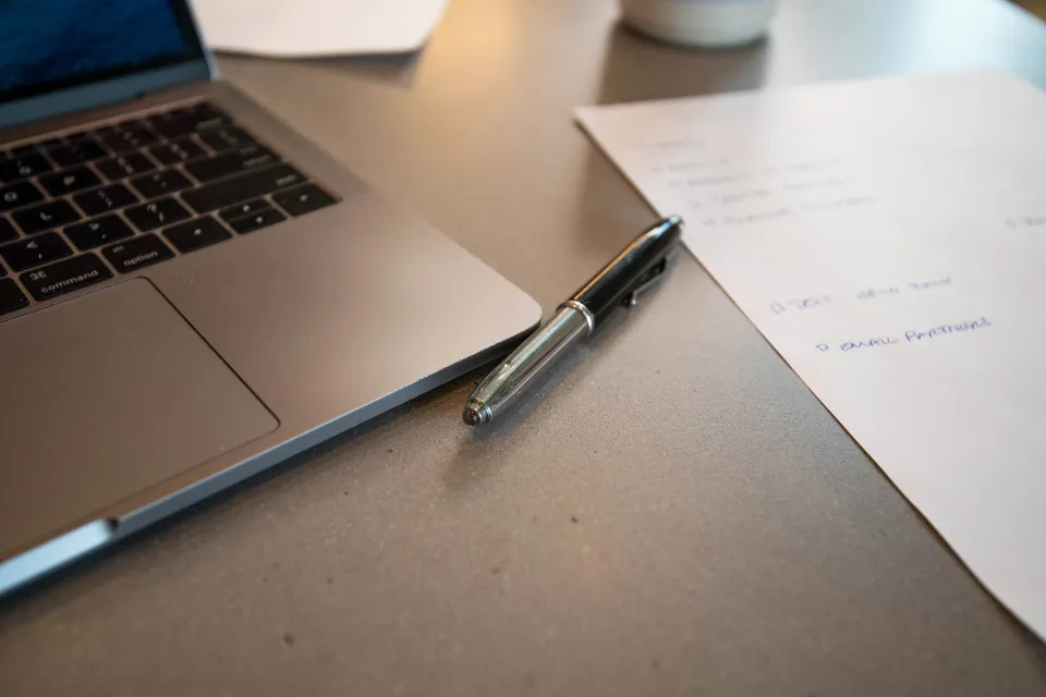 A laptop on a desk next to a pen and handwritten notes